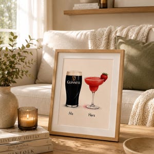 May include: Framed print featuring a pint of Guinness and a red cocktail with a strawberry garnish. The words "His" and "Hers" are below the drinks. The frame is light brown and sits on a wooden table.