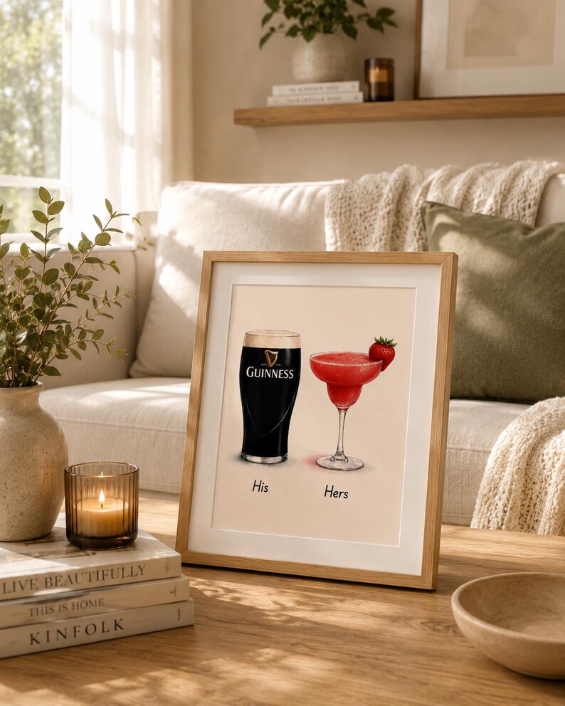 May include: Framed print featuring a pint of Guinness and a red cocktail with a strawberry garnish. The words "His" and "Hers" are below the drinks. The frame is light brown and sits on a wooden table.