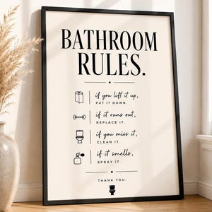 May include: A framed wall art print with a cream background and black text reading BATHROOM RULES. It features four icons with instructions: if you lift it up, PUT IT DOWN; if it runs out, REPLACE IT; if you miss it, CLEAN IT; if it smells, SPRAY IT.