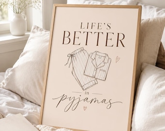 May include: A framed print with the text "LIFE'S BETTER in pyjamas" in a stylish font. The print features a sketch of pajamas. The frame is a light brown color. The print is on a bed with pillows and a blanket.