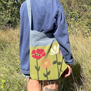 Può includere: Borsa a tracolla all'uncinetto fatta a mano con cinturino e sezione superiore azzurra. La sezione inferiore della borsa è verde oliva, con disegni di fiori rossi, rosa e bianchi. La borsa è indossata sopra una camicia blu.