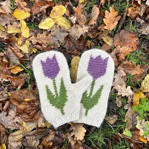 May include: Off-white crochet mittens featuring a purple tulip design with green stems and leaves. The mittens are displayed on a bed of autumn leaves and grass.