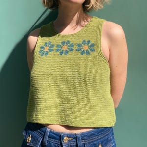 以下が含まれることがあります： 胸に青と黄色の花の模様が3つあしらわれた、グリーンのクロシェタンクトップ。ノースリーブで、丸いネックラインです。モデルは青いデニムジーンズを着用しています。