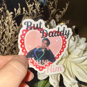 May include: A sticker with the text "But Daddy Love Him" in a heart shape. The sticker features a photo of a person and a holographic heart. The sticker is held in front of a bouquet of flowers.