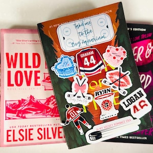 May include: Three books with colorful covers and stickers. The central book has hockey-themed stickers, including a jersey with "Graham 44." The other books are pink and black, with titles like "Wild Love" and "Forever & Always."