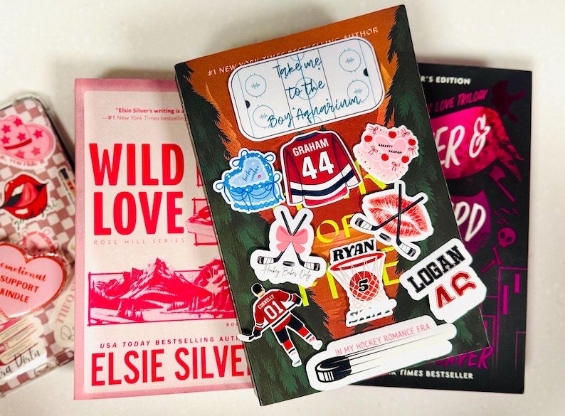May include: Three books with colorful covers and stickers. The central book has hockey-themed stickers, including a jersey with "Graham 44." The other books are pink and black, with titles like "Wild Love" and "Forever & Always."