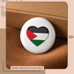 Può includere: Un bottone bianco con un design a forma di cuore con i colori della bandiera palestinese: rosso, nero, bianco e verde. Il bottone è posto su uno sfondo marrone, con il testo "MORE DESIGNS AVAILABLE" in basso.