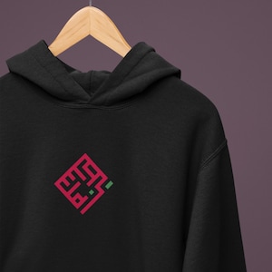 Pode incluir: Camisola com capuz preta com um design geométrico vermelho e verde na frente. O design é um padrão angular estilizado. A camisola está pendurada num cabide de madeira contra um fundo roxo.