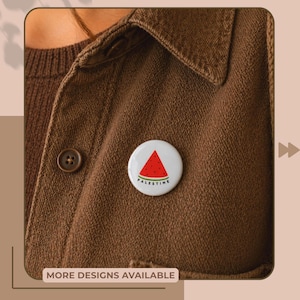 May include: A white button pin with a watermelon slice design and the word "PALESTINE" in green. The pin is attached to a brown jacket. The text "MORE DESIGNS AVAILABLE" is at the bottom of the image.