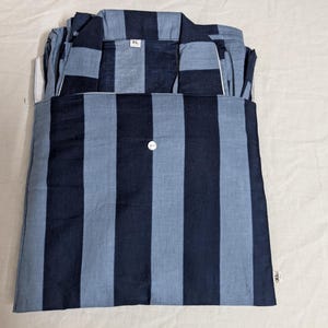 Puede incluir: Una parte superior de pijama doblada con un patrón de rayas verticales en azul marino oscuro y azul claro. La prenda tiene un cuello, un único botón blanco centrado y una etiqueta de talla blanca con la inscripción XL visible en el cuello y la costura lateral.