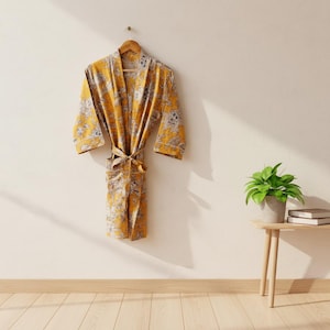 Puede incluir: Una bata amarilla con estampado floral cuelga de una percha de madera contra una pared color crema. La bata tiene una cintura anudada y ribetes blancos. Una pequeña mesa auxiliar tiene una planta en maceta y libros.