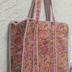 May include: A floral print tote bag with a beige background and orange and pink flowers. The bag has matching handles and trim with a striped pattern. The bag is rectangular and appears to be quilted.