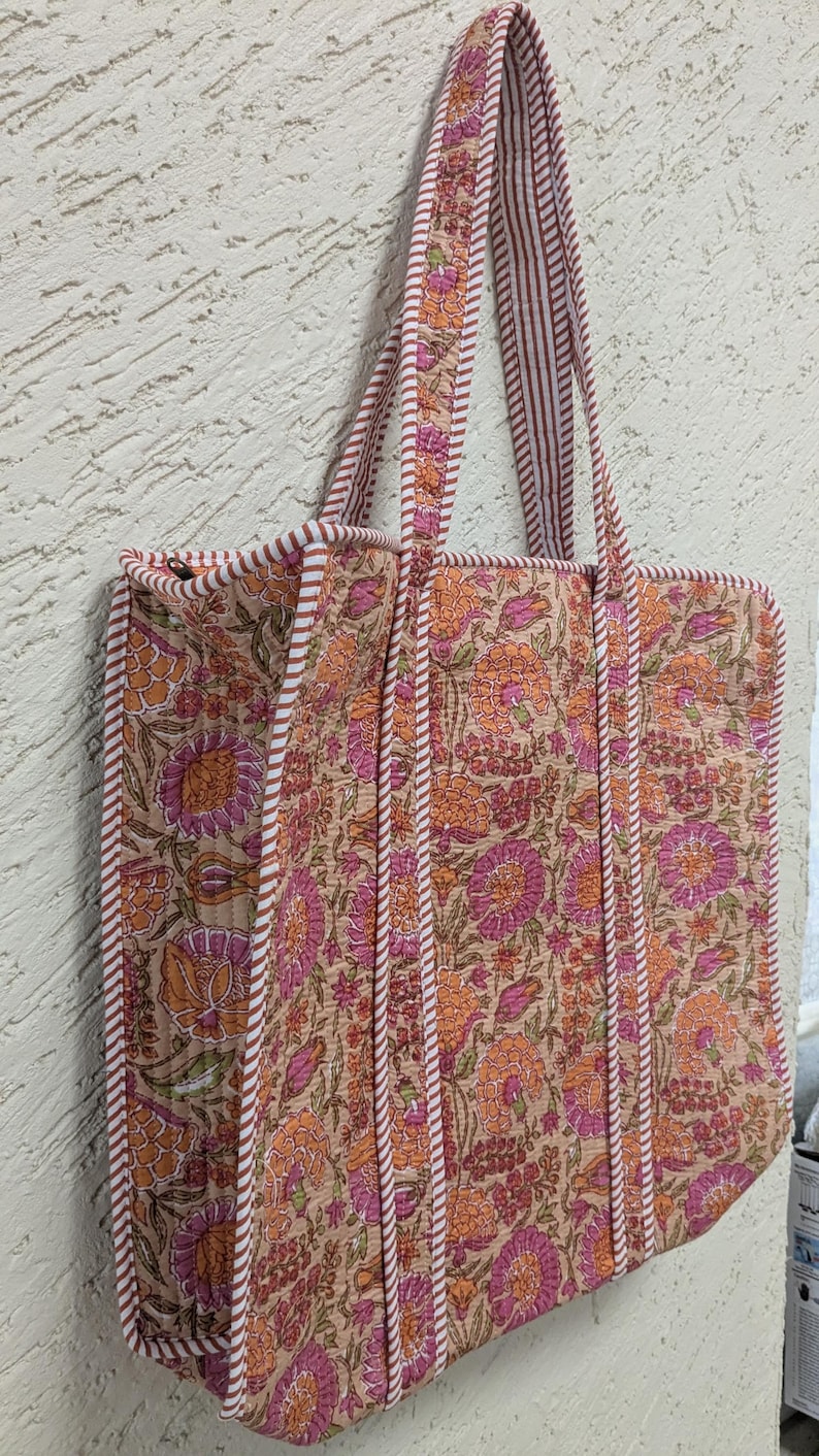 May include: A floral print tote bag with a beige background and orange and pink flowers. The bag has matching handles and trim with a striped pattern. The bag is rectangular and appears to be quilted.