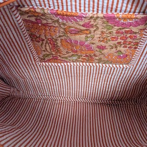 May include: Interior view of a tote bag. The inside features a brown and white striped fabric. A pocket is visible with a floral pattern in shades of orange, pink, and green. The bag's trim is a solid orange color.