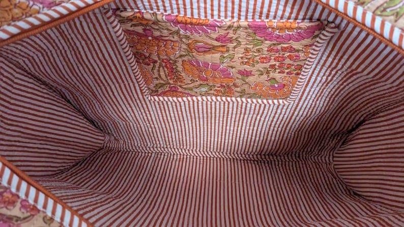 May include: Interior view of a tote bag. The inside features a brown and white striped fabric. A pocket is visible with a floral pattern in shades of orange, pink, and green. The bag's trim is a solid orange color.