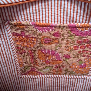 May include: Interior view of a tote bag with a floral pocket. The bag's lining features a white and brown striped pattern. The pocket showcases a detailed floral design in shades of pink, orange, and beige.
