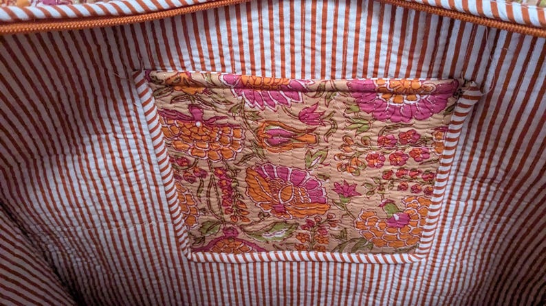 May include: Interior view of a tote bag with a floral pocket. The bag's lining features a white and brown striped pattern. The pocket showcases a detailed floral design in shades of pink, orange, and beige.