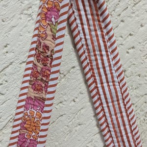 May include: Close-up of a quilted fabric strap with a floral pattern and red and white stripes. The floral section features pink and orange flowers on a beige background. The striped section has red stripes.