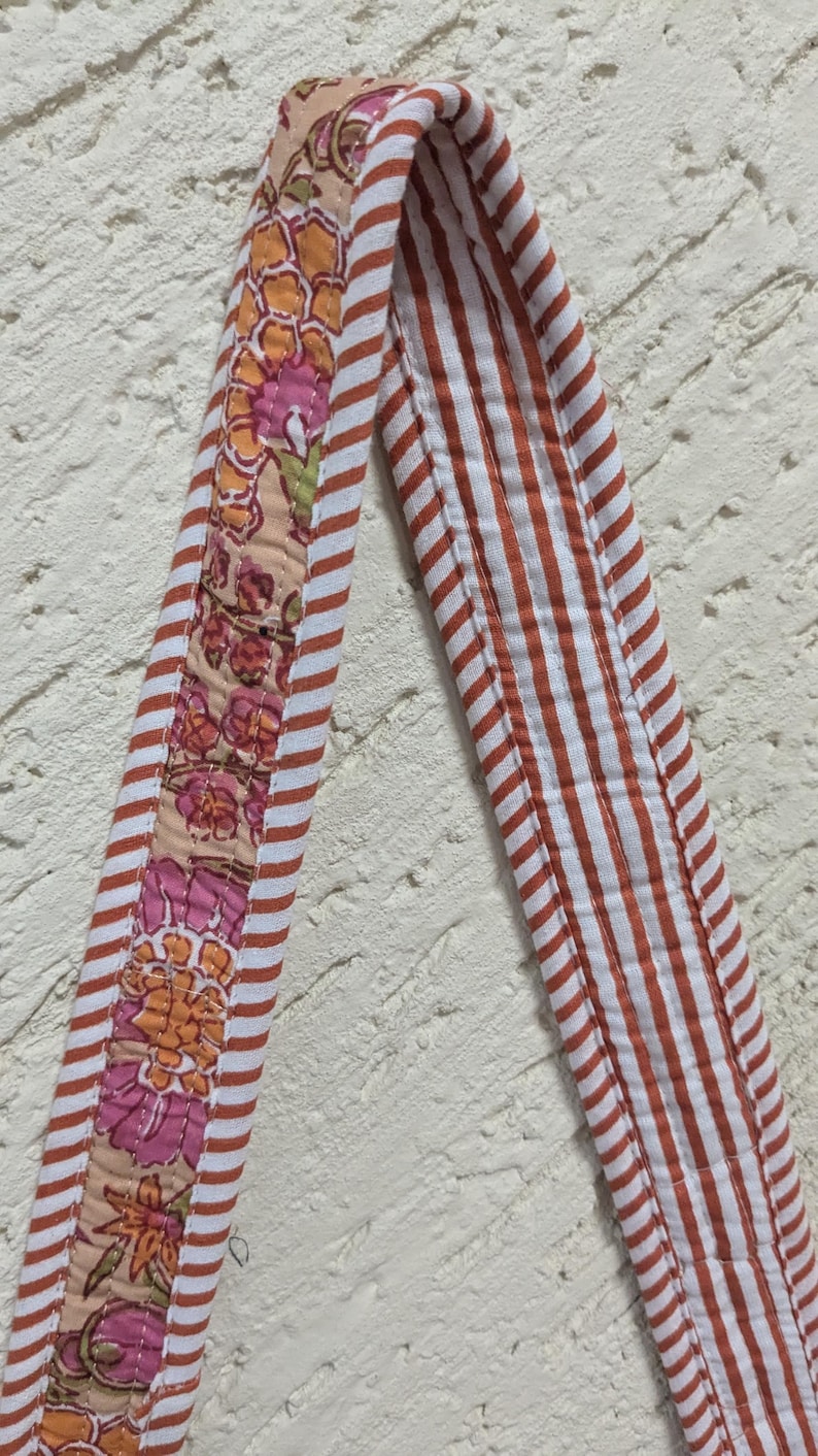 May include: Close-up of a quilted fabric strap with a floral pattern and red and white stripes. The floral section features pink and orange flowers on a beige background. The striped section has red stripes.