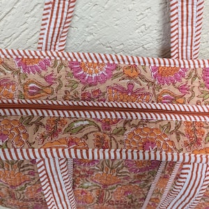 May include: Close-up of a floral print tote bag. The bag features a beige background with orange and pink floral patterns. The bag has a brown zipper and trim, and white and brown striped handles.