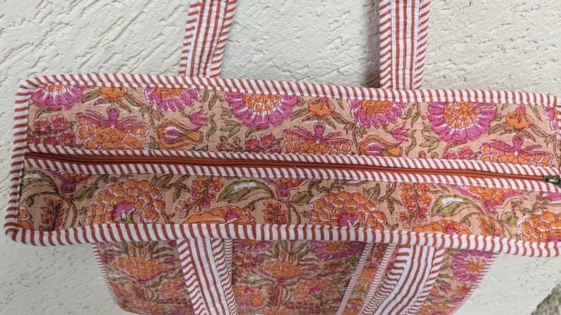 May include: Close-up of a floral print tote bag. The bag features a beige background with orange and pink floral patterns. The bag has a brown zipper and trim, and white and brown striped handles.