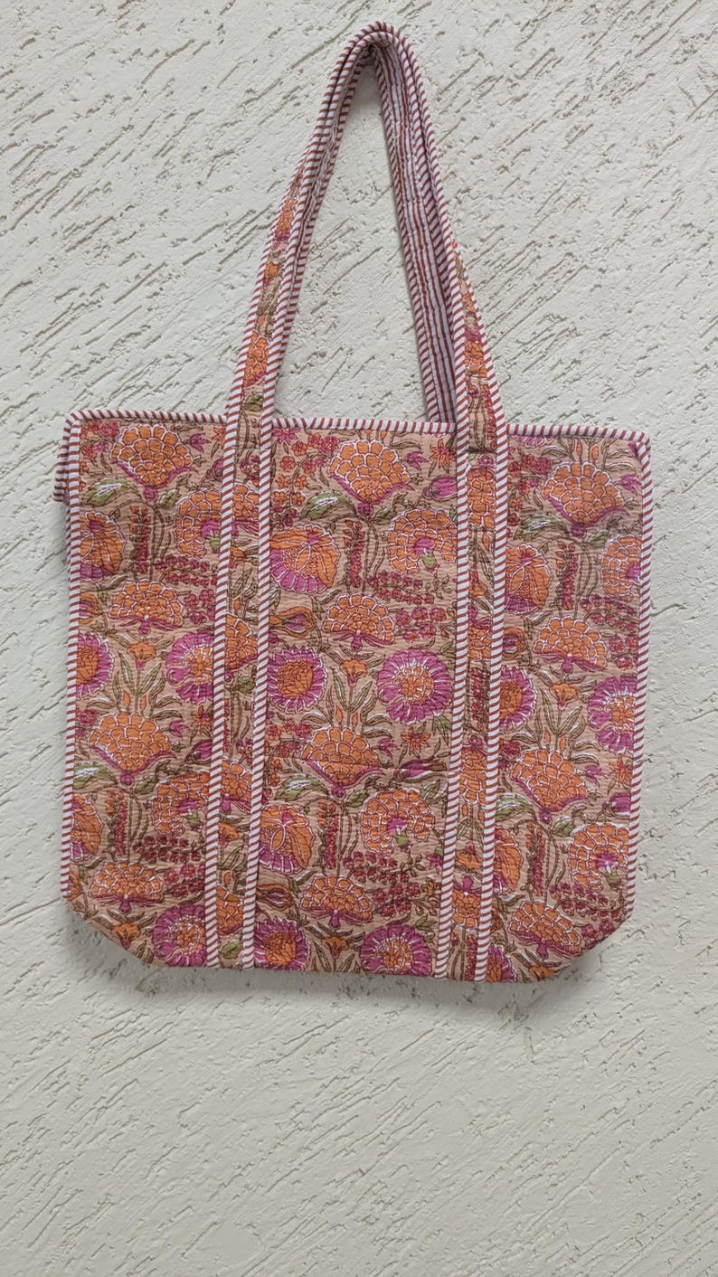 May include: A floral tote bag with a beige background and orange and pink flower pattern. The bag has matching handles and trim with a striped design. The bag is rectangular and appears to be made of fabric.