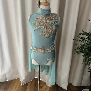 May include: A light blue leotard featuring a high neckline, embellished with intricate floral lace appliques, clear crystals, and beads. The garment includes a matching beaded belt at the waist and a sheer, flowing side panel attached to the hip.