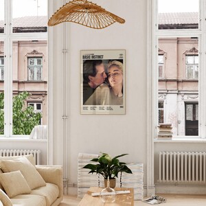 May include: A framed movie poster for "Basic Instinct" featuring a close-up of two people. The poster is in a room with a beige sofa, wooden table, and a woven light fixture. The poster is in a gold frame.