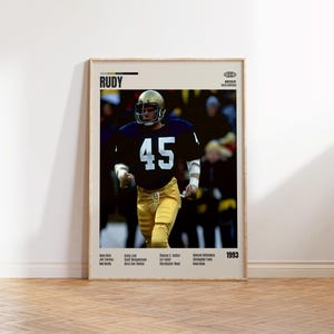 May include: Framed poster featuring a football player in a navy blue jersey with the number 45, gold pants, and a gold helmet. The poster includes the title "RUDY" and the year 1993, along with a list of names. The background is a blurred image of a crowd.