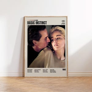 Może przedstawiać: Oprawiony plakat filmowy do filmu "Basic Instinct" z Michaelem Douglasem i Sharon Stone. Plakat ma beżowe tło z tytułem filmu pogrubionymi czarnymi literami. Rok 1992 jest wyświetlany na dole.