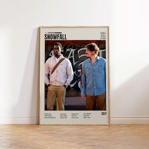 May include: A framed poster for the TV show "Snowfall" featuring two men in front of graffiti. The poster has a beige frame and includes the show's title, cast names, and the year 2017. The background is a white wall and a wooden floor.
