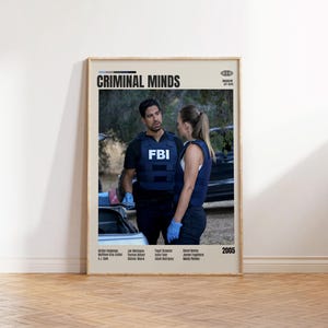 May include: A framed poster for the TV show "Criminal Minds" featuring two people in tactical vests with the FBI logo. The poster has the show's title at the top and the year 2005 at the bottom. The poster is displayed on a wooden floor.