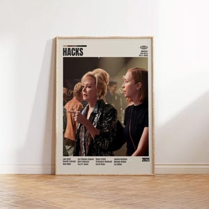Op de afbeelding: Een ingelijste poster voor de tv-show "Hacks" met twee vrouwen in een scène. De poster heeft een beige achtergrond met de titel van de show bovenaan en het jaar 2021 onderaan. De poster wordt op een witte muur weergegeven.