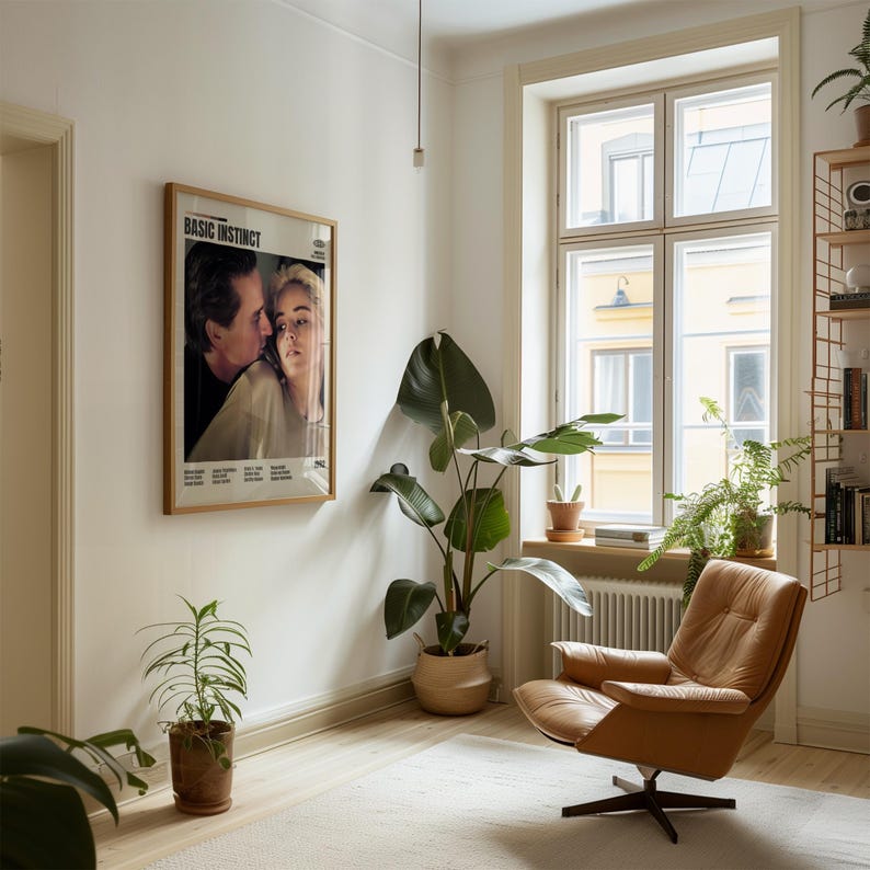 May include: A living room scene featuring a framed "Basic Instinct" movie poster, a tan leather armchair, and several potted plants. The room has a neutral color palette with a large window and a light-colored rug.