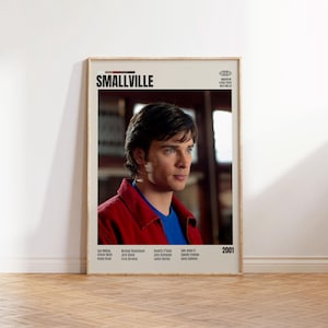 以下が含まれることがあります： 「SMALLVILLE」のタイトルが黒で表示された額入りのポスター。赤いジャケットと青いシャツを着た男性が描かれています。ポスターには2001年と名前のリストが含まれています。背景はぼやけたインテリアです。