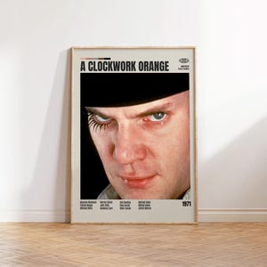 May include: A framed movie poster for "A Clockwork Orange" featuring a close-up of a man's face with a black hat and exaggerated eyelashes. The poster has the film's title and the year 1971.