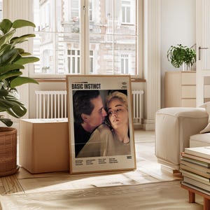 May include: A framed movie poster for "Basic Instinct" featuring Michael Douglas and Sharon Stone. The poster has a beige border and is displayed in a well-lit room with a large window and a wooden floor. The poster is dated 1992.