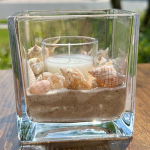 May include: A square glass candle holder filled with sand, seashells, and a white candle. The seashells are various sizes and colors, including tan, white, and orange. The candle is in a clear glass holder.