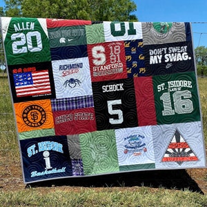 May include: A green, red, blue, and white patchwork quilt made from old t-shirts. The quilt is hanging on a fence with a green grassy field in the background. The t-shirts have various logos and text, including "Allen 20", "Richmond Spiders", "Stanford Cardinal", "Schhock 5", "Don't Sweat My Swag", "St. Isidore 16", and "Chico State".