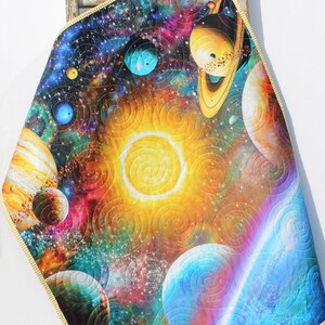May include: A colorful space-themed quilt with a yellow sun, blue planets, and swirling nebulae. The quilt is quilted with a spiral pattern.