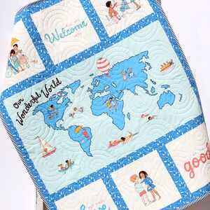 May include: A blue and white quilted baby blanket with a world map design. The map is surrounded by a blue border and has a hot air balloon, sailboat, and children playing on it. The blanket has several squares with text, including "Our Wonderful World", "Welcome", "Love", and "Be the good".
