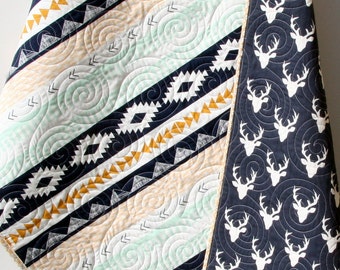 May include: A baby quilt with a geometric pattern in black, white, gold, and mint green. One side of the quilt features a repeating pattern of white deer silhouettes on a navy blue background.