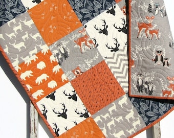 May include: A patchwork baby quilt with a woodland theme. The quilt is made of various fabrics in shades of gray, orange, and white. The fabric patterns include deer, bears, and foxes.