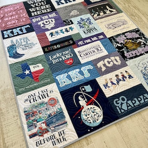 以下が含まれることがあります： 様々なTシャツの正方形で構成された手作りのキルトで、テキストとグラフィックが特徴です。キルトは青、紫、白など、さまざまな色を示しています。「Wish You Were Here」や「Out of This World」などのフレーズが含まれています。