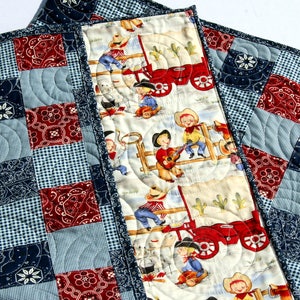 May include: A patchwork quilt with a blue and red checkered pattern and a bandana print. The back of the quilt features a white fabric with a repeating pattern of cowboys riding horses and driving a red wagon.