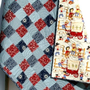 May include: A red, white, and blue patchwork quilt with a bandana print. The back of the quilt features a pattern of cowboys and a wagon.