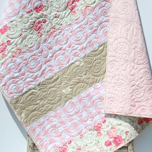 May include: A pink and brown patchwork quilt with a floral pattern and a swirl design. The quilt is folded in half and is lying on a white surface.