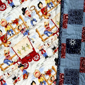 May include: A white quilt with a patchwork design featuring cowboys and a wagon. The quilt is made of cotton fabric and has a blue and red checkered backing.