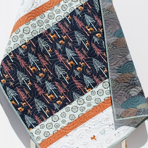 May include: A baby quilt with a woodland theme. The front features a navy blue fabric with a pattern of trees and deer. The back features a white fabric with a pattern of foxes. The quilt is trimmed with a teal border.