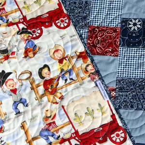 May include: A white and cream quilt with a patchwork design featuring red, blue, and white gingham patterns and blue bandana prints. The quilt is quilted with a swirl pattern and features a scene of cowboys on a wagon.